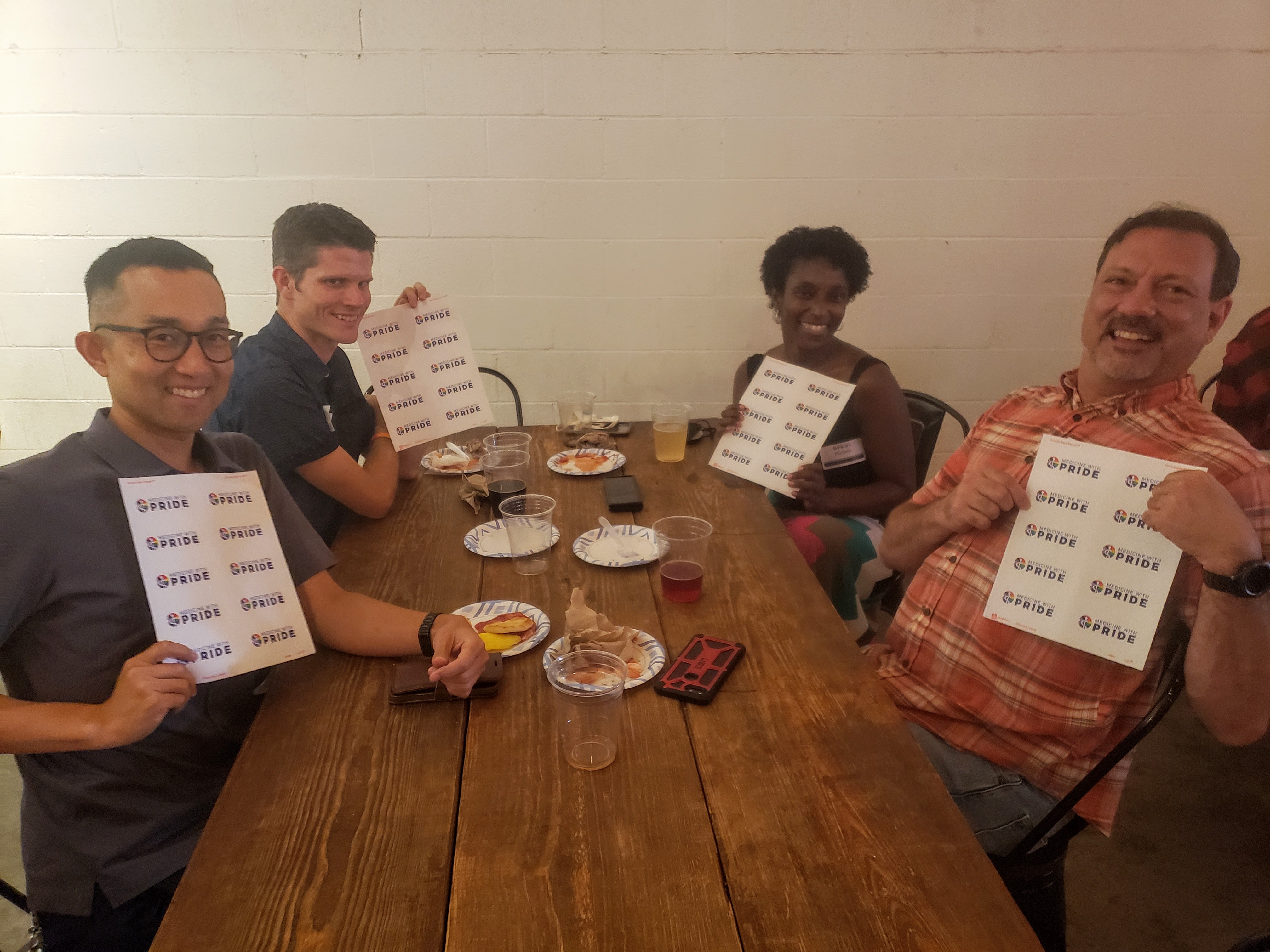 Four people sit at a table holding up pride in medicine stickers and smiling.