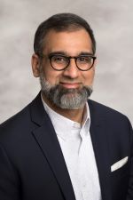 faintly smiling man with salt and pepper full beard and short dark hair wearking round glasses and a dark suit jacket over a white shirt