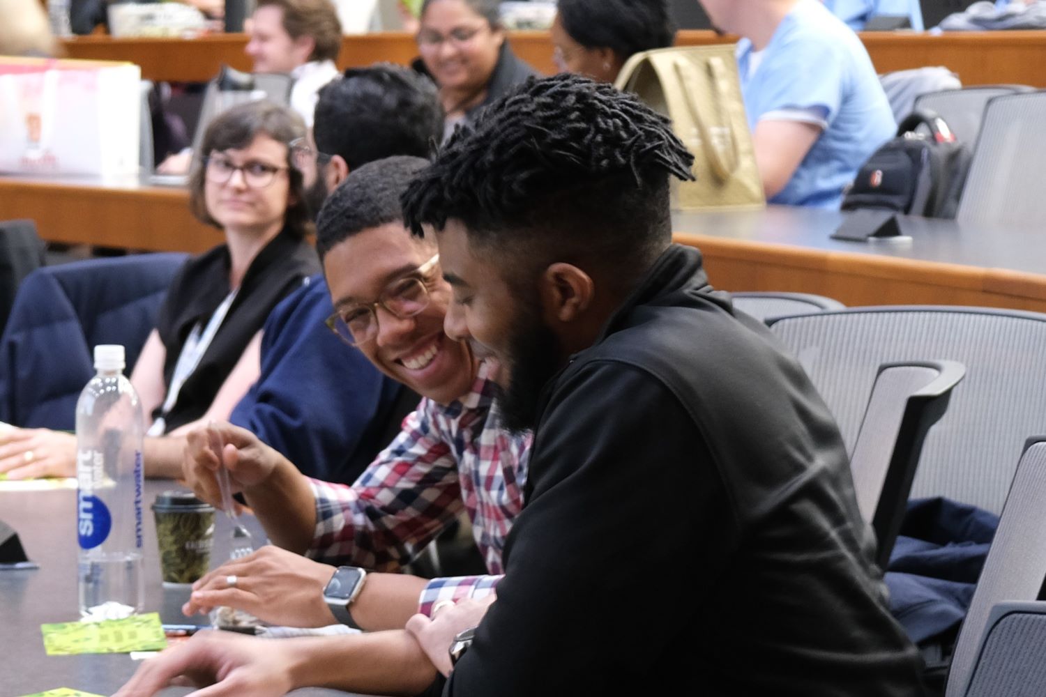 two people laughing and talking over lunch in a large lecture hall with others in the background