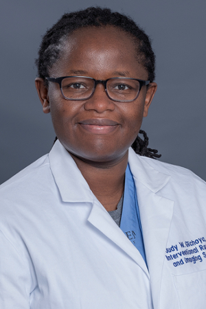 smiling woman with black hair pulled back into short braids wearing black rimmed spectacles and a white doctor coat over a light blue shirt