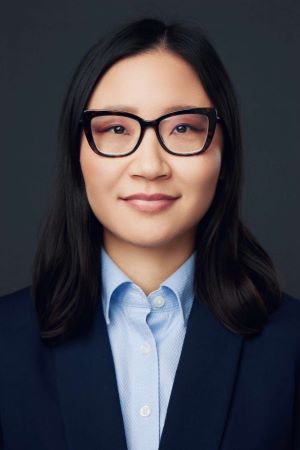 faintly smiling person wearing black rimmed spectacles and a navy blazer over a light blue blouse