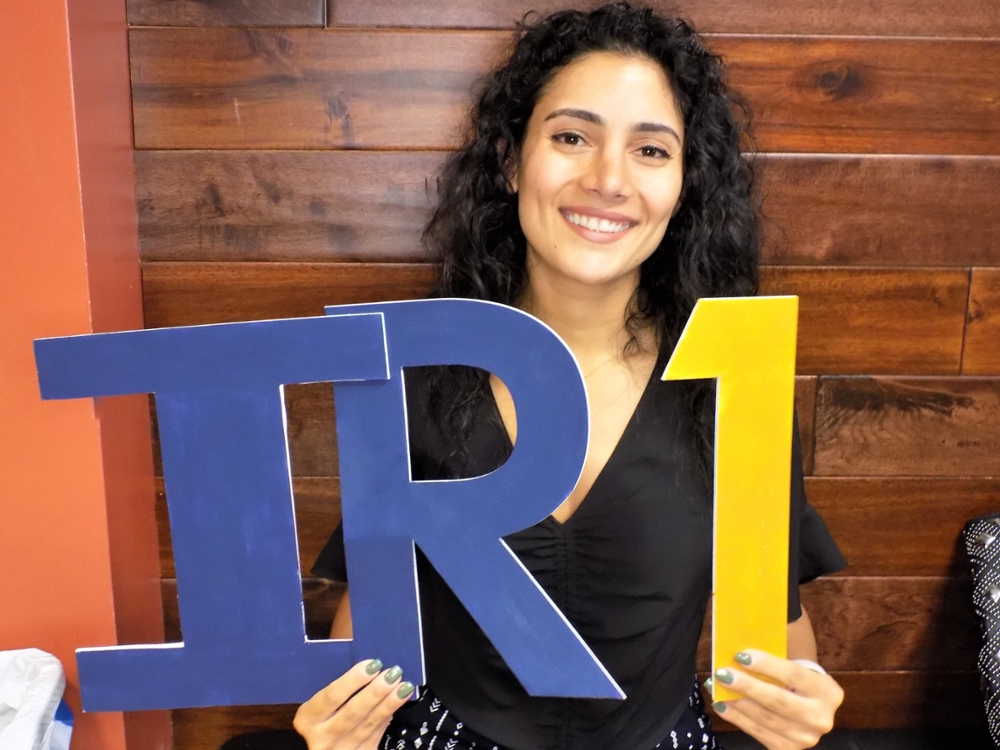 smiling woman with curly dark long hair holding blue letters i and r and gold number 1