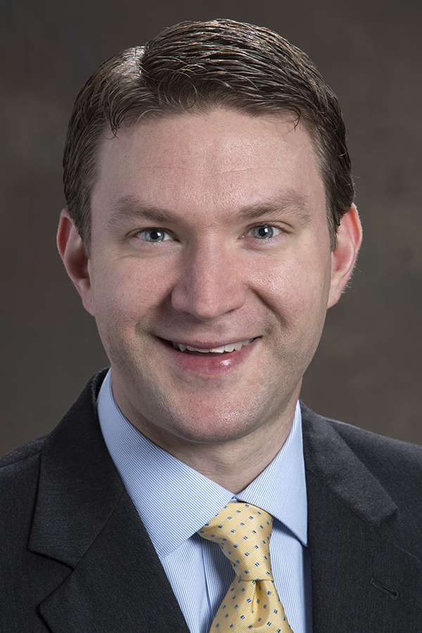 smiling man with short brown hair and wearing a dark jacket gold tie and light blue button down shirt